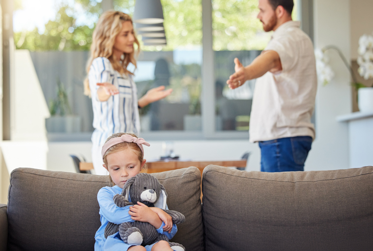 Image of sad girl sitting on couch with parents arguing behind her.