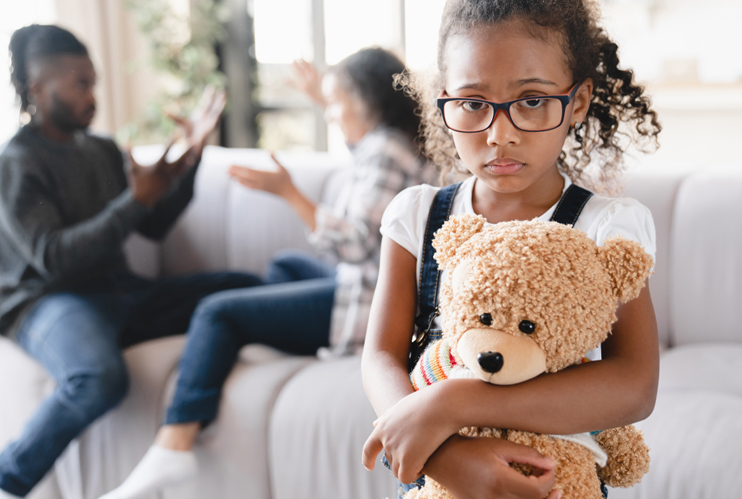Image of sad girl holding teddy bear with parents in the background arguing.